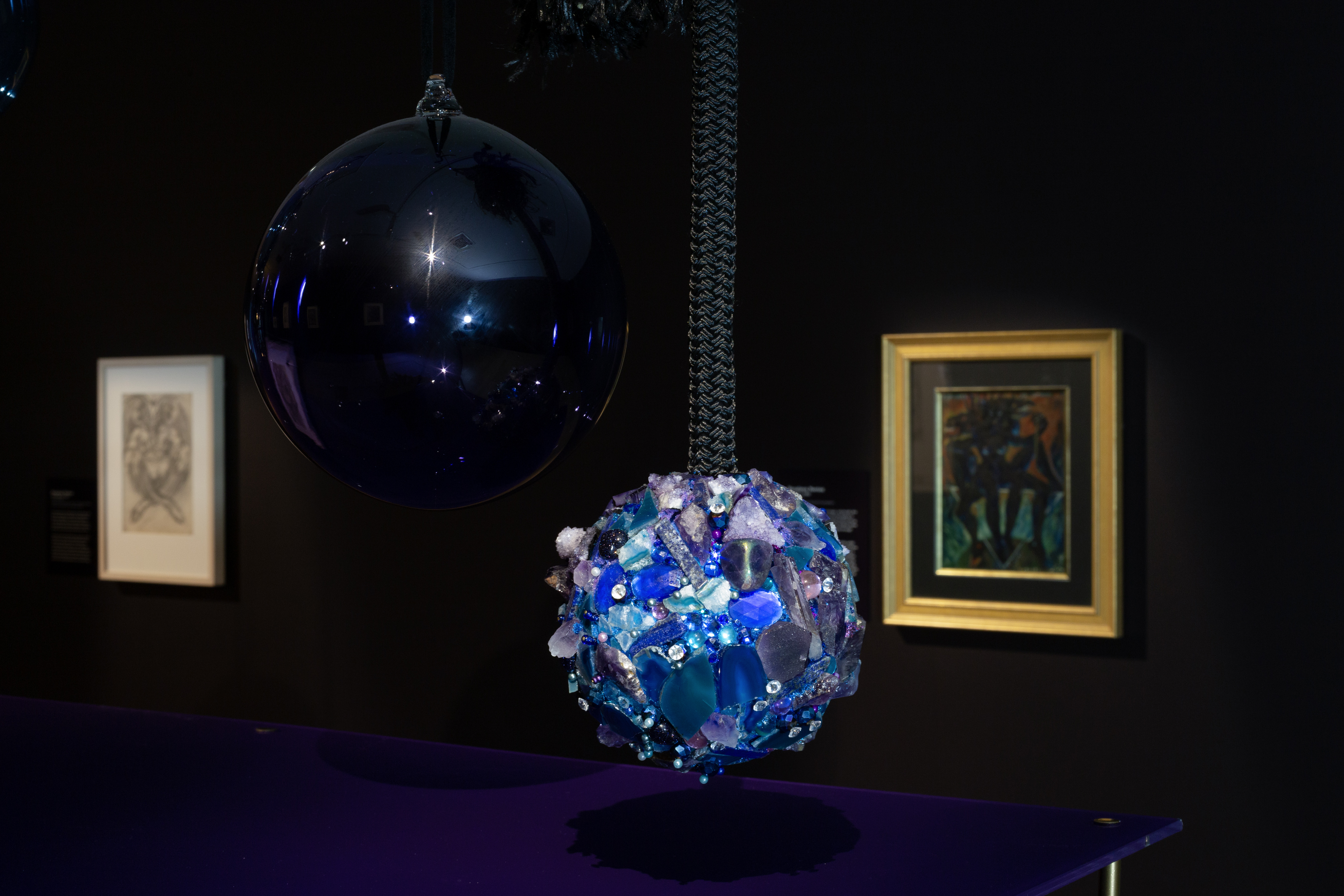 Image of two hanging glass orbs, with exhibition paintings in the background. 