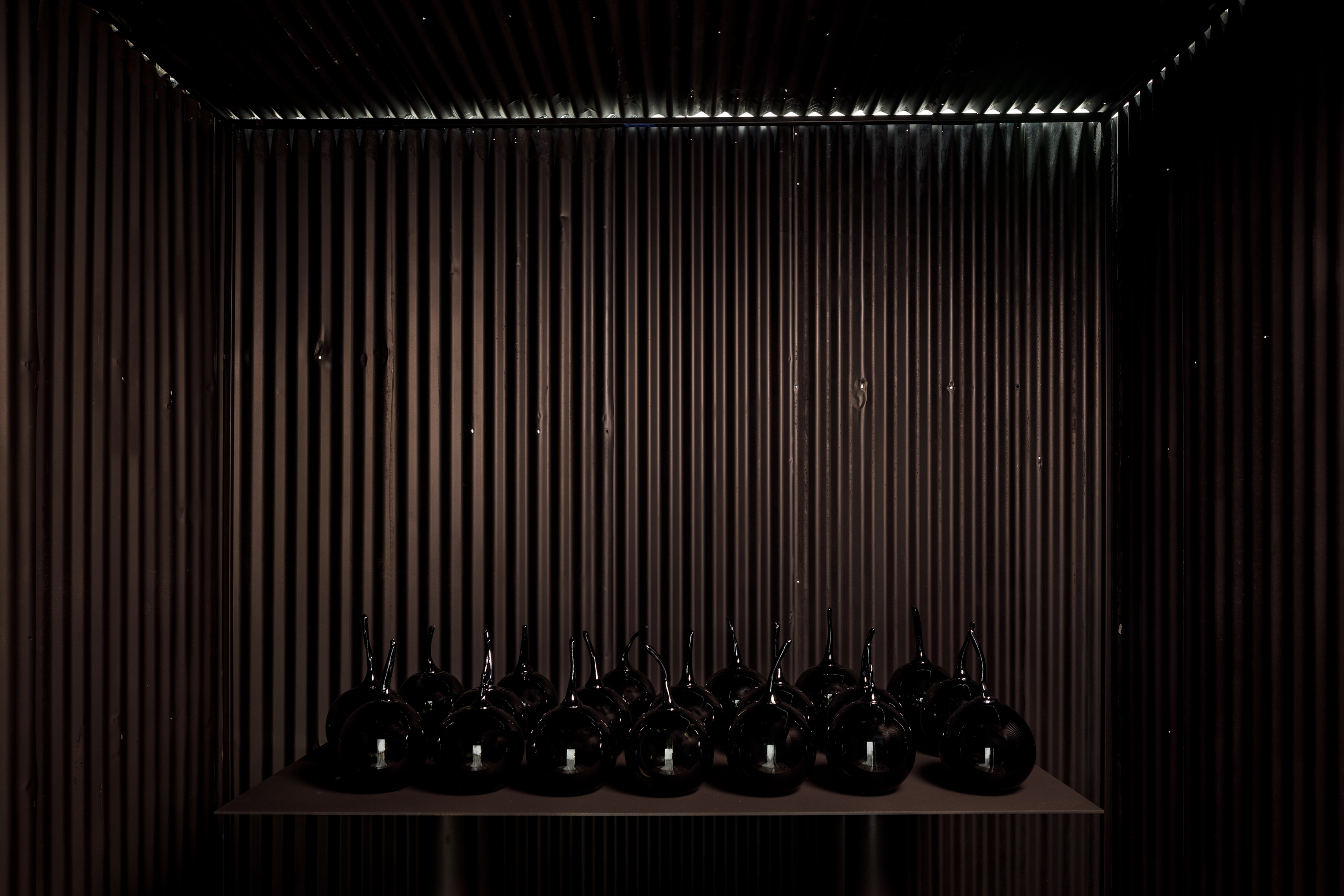 Installation view of the inside of one of Yhonnie Scarce’s corrugated Zincalume sheds, or structures in the work Missile Park. You can only see inside the dark shed, the glass work is visible on the table. Taken at the Australian Centre for Contemporary Art.