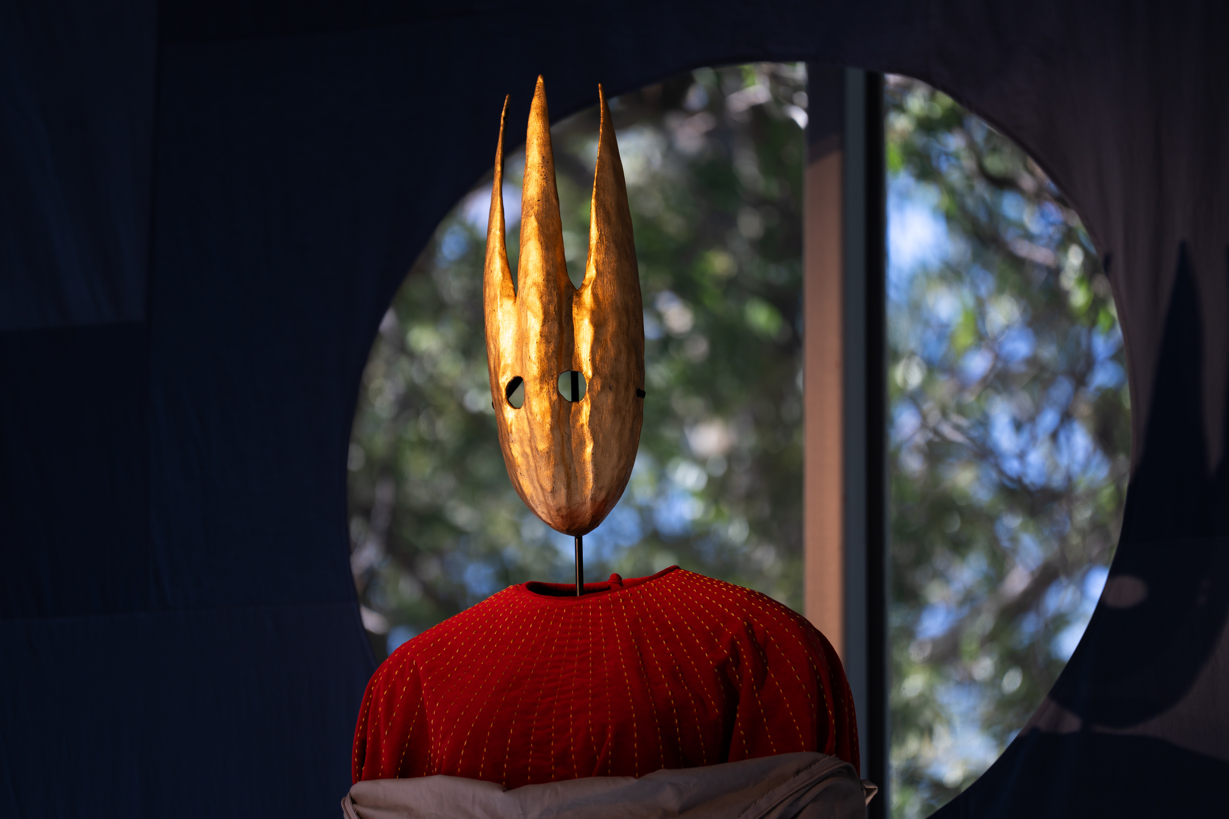 Golden mask against a blue background with circular opening looking out to trees.