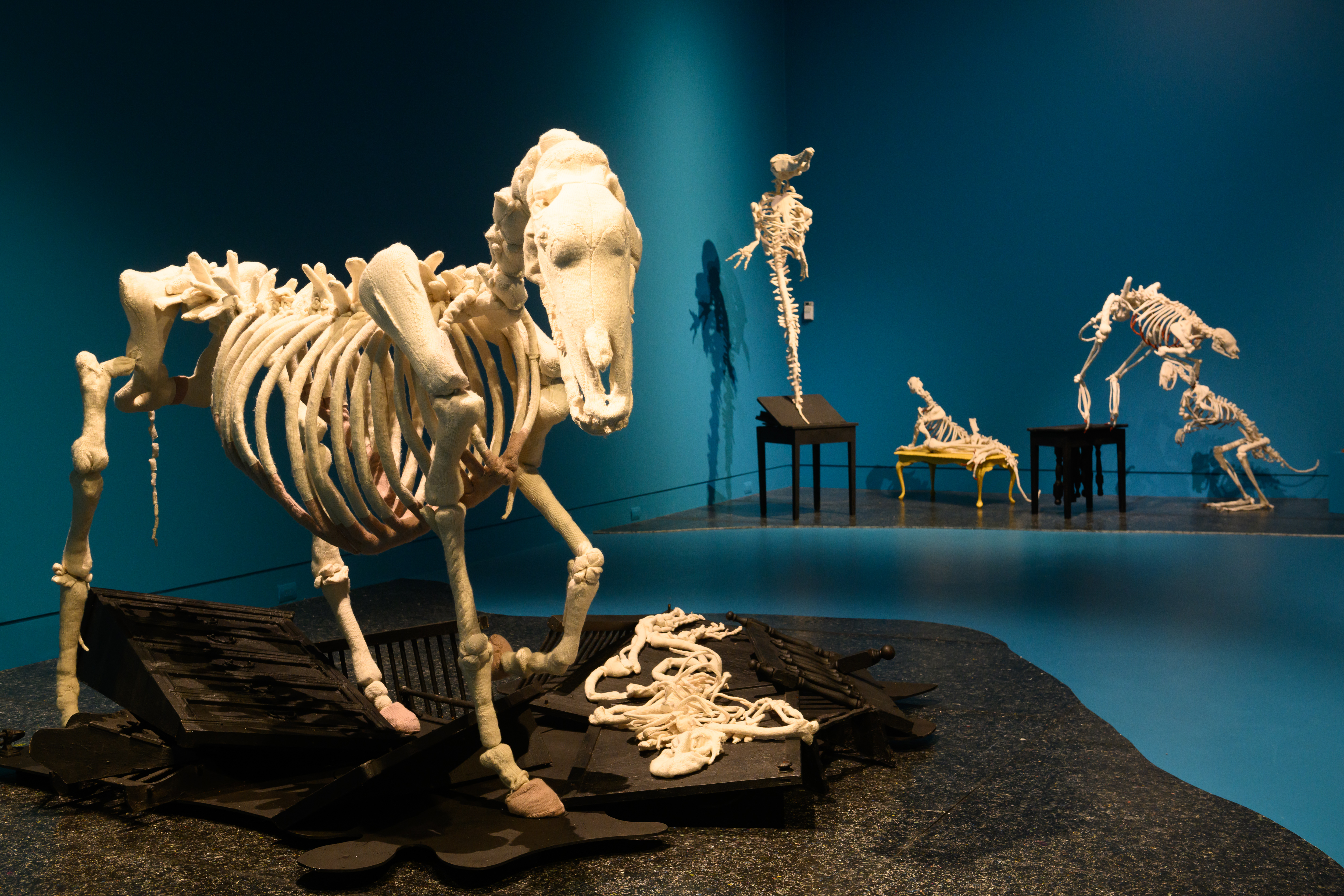 A life-sized skeleton of a horse knitted by Michele Beevors. Its hoof is raised above a limp knitted skeleton of a human. Both surrounded by second-hand furniture that has been painted with tar paint. The floors of the Gallery are painted a deep ocean blue and the artworks are on a composite black board.
