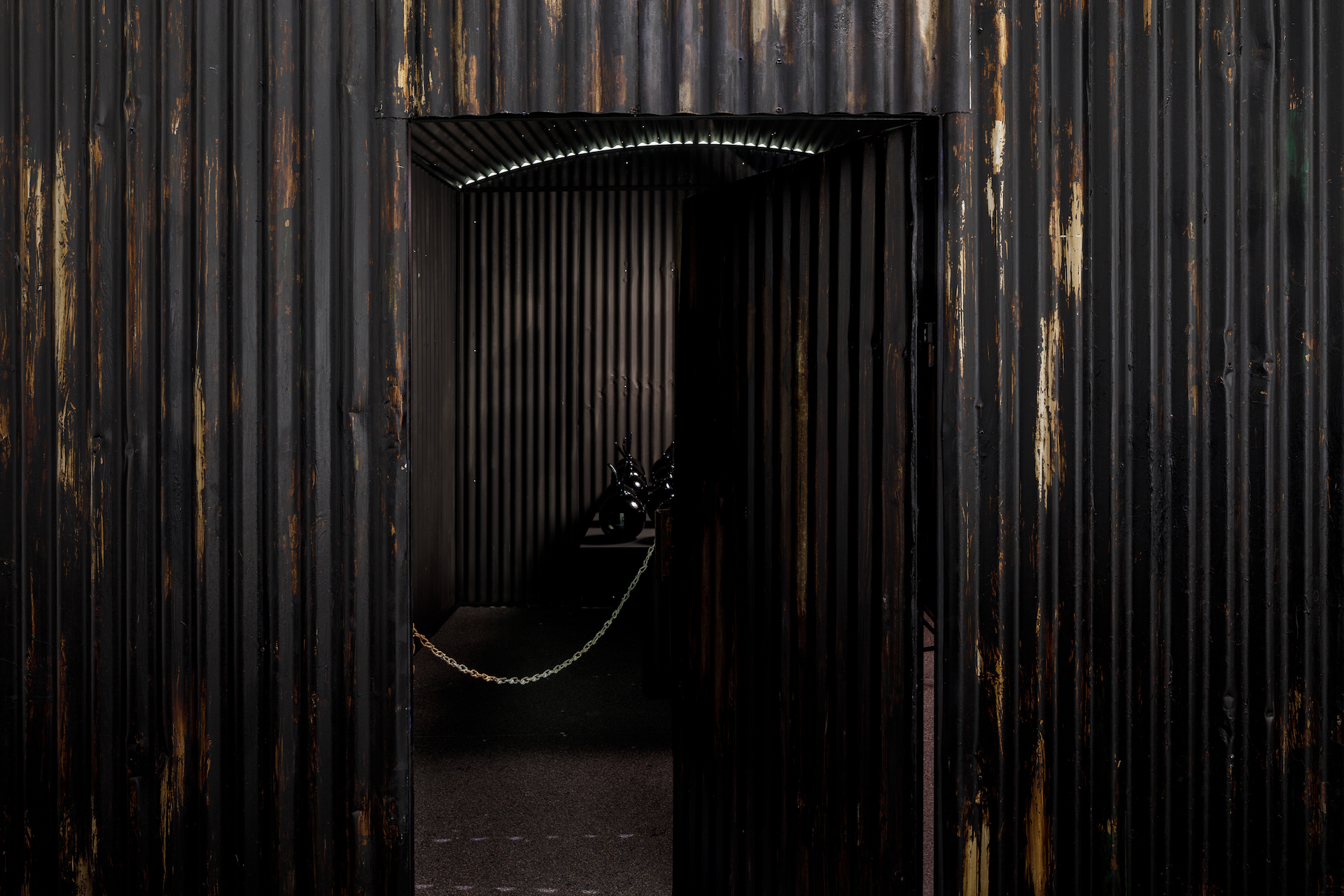 Installation view of the entrance to one of Yhonnie Scarce’s corrugated Zincalume sheds, or structures in the work Missile Park. Inside the dark shed, the glass work is visible on the table. Taken at the Australian Centre for Contemporary Art.